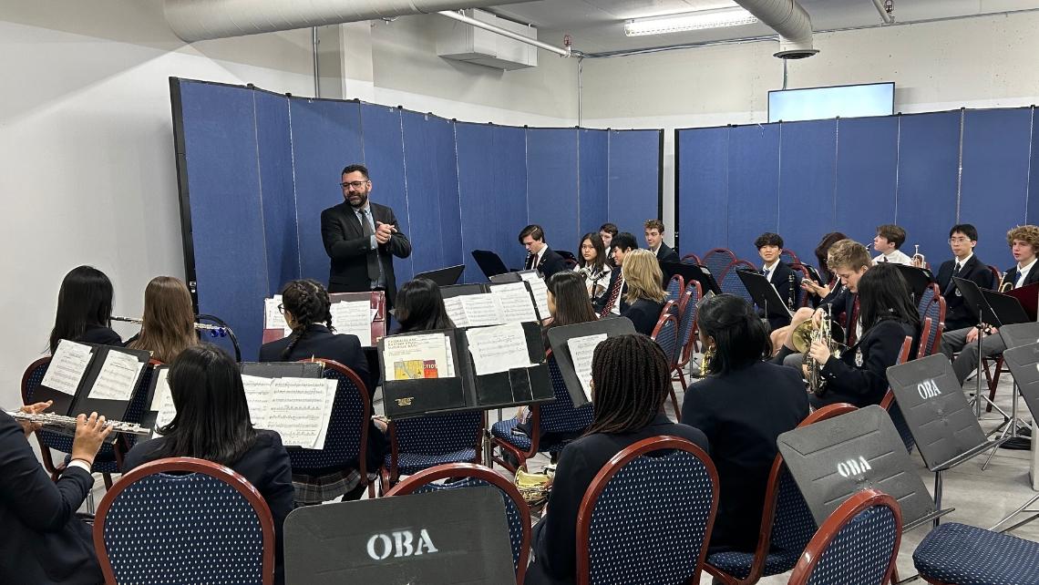 A student band, seat in front of music stands, faces a conductor in front of a blue curtain