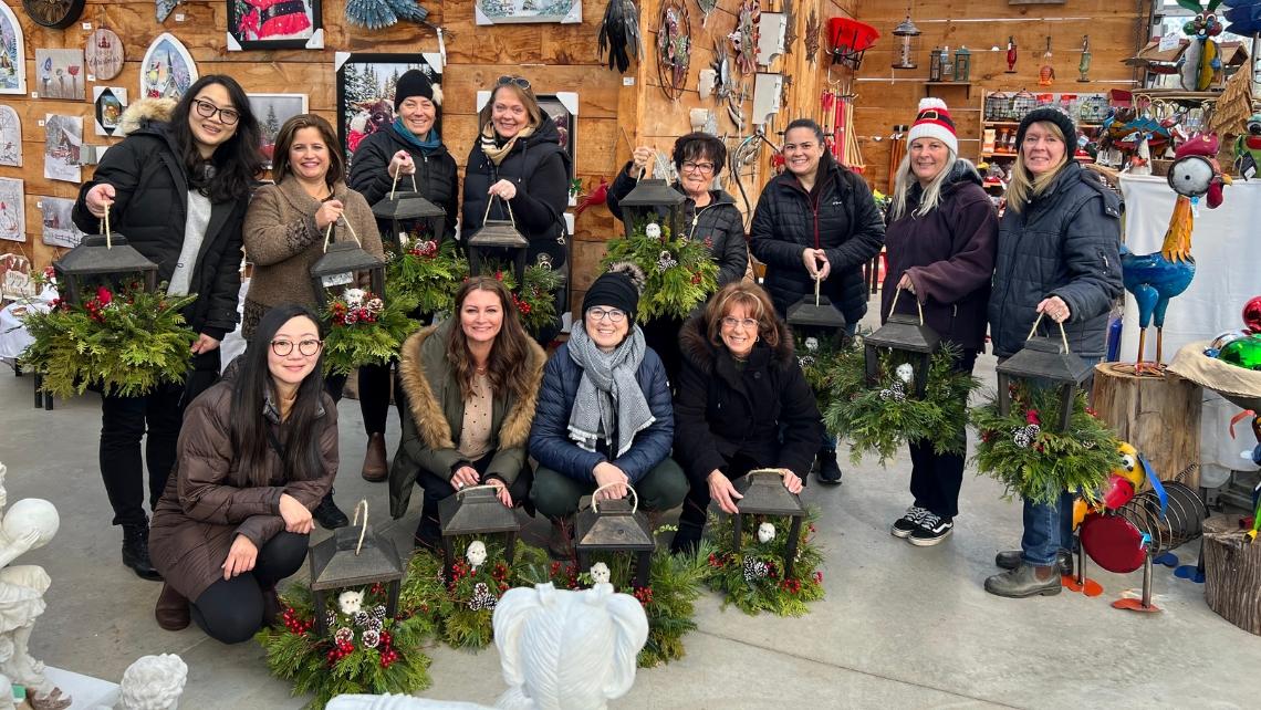 A group of adults, some standing and some kneeling, and they are each holding a black metal lantern decorated with evergreens, red berries and pinecones