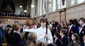 Sacristan carrying a large cross up the aisle of a chapel filled with students wearing uniforms with poppies
